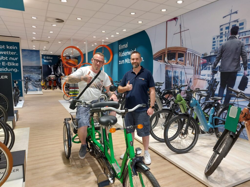 Im Verkaufsraum sitzt Andreas auf einem grünen Dreiradtandem er strahlt in die Kamera und zeigt einen Daumen hoch. Rechts neben ihm steht Florian, ein Mitarbeiter von Küstenrad und schaut in die Kamera und hält auch einen Daumen hoch.